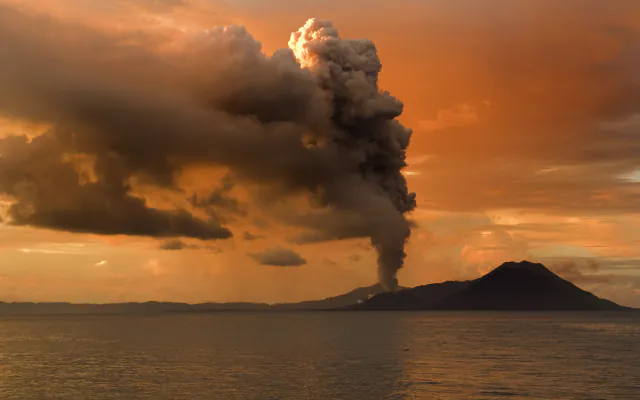 A grey ash column rising from a low volcanic ridge across calm water under an orange sunset sky, with a darker cone on the shoreline to the right