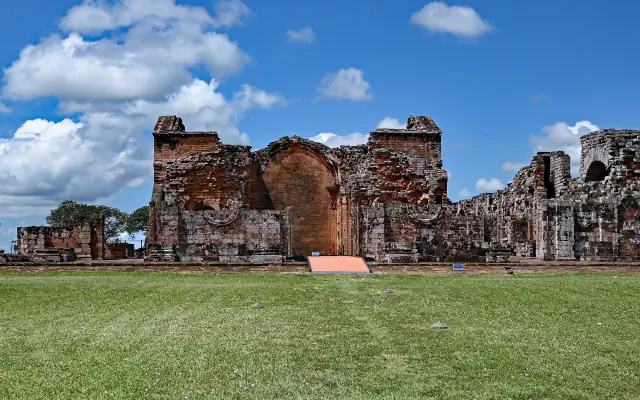 Ruined red-brick and stone walls of the main church at La Santísima Trinidad de Paraná behind a wide lawn, with a tall central arch under a blue sky and white clouds