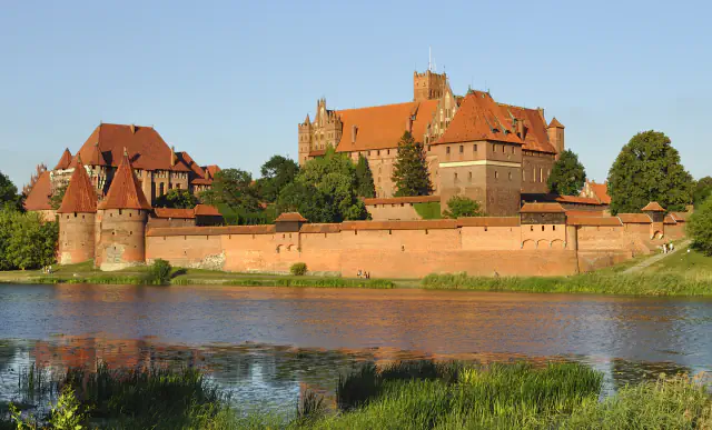 Red-brick Malbork Castle across the Nogat river in warm low sunlight: a long curtain wall with round towers at the left and the High Castle with its tower rising behind