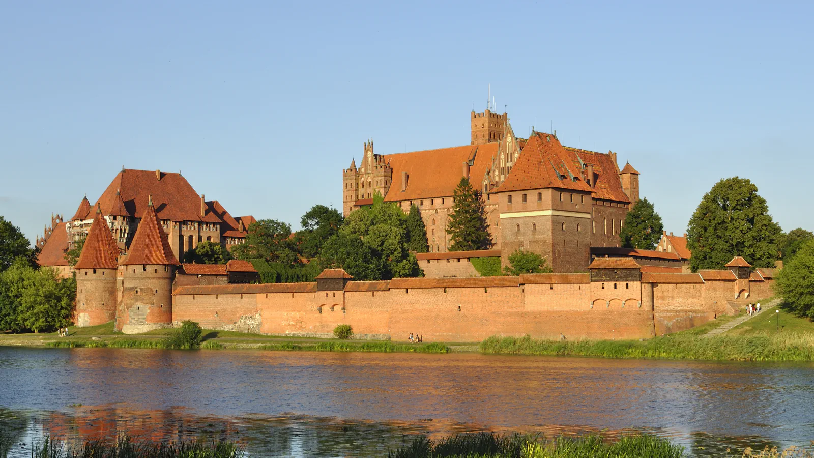 Grad Malbork iz rdeče opeke ob reki Nogat v topli svetlobi nizkega sonca: dolgo obzidje z okroglimi stolpi na levi in Visoki grad s stolpom, ki se dviga za njim