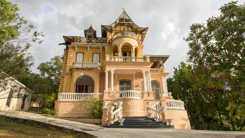 Villa Miramar v četrti Pacot, pogled z dovoza: rumeno-kremna hiša gingerbread s stolpičasto zgornjo etažo, balkoni in belim stopniščem z balustrado, obdana z drevjem pod oblačnim nebom