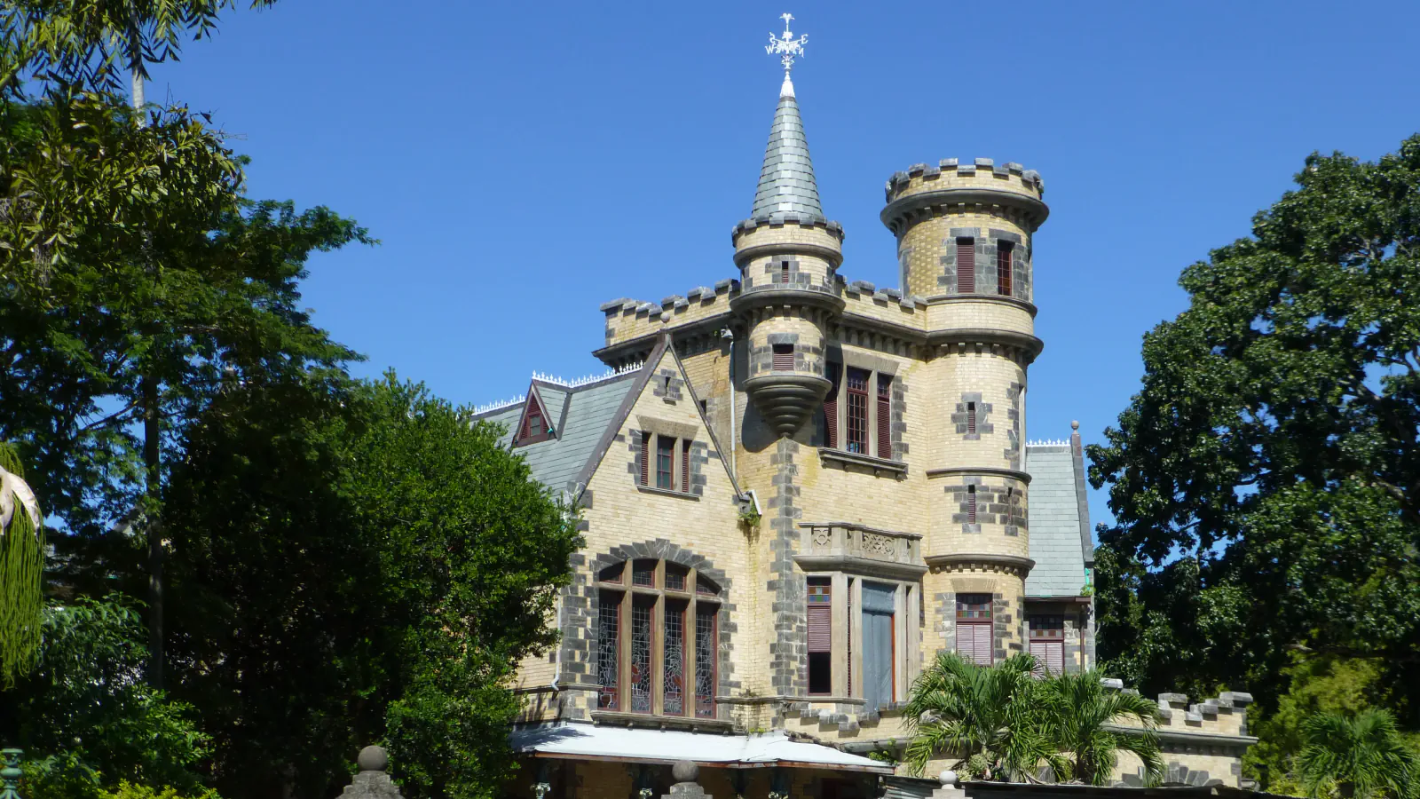 Kamnita stavba Stollmeyer’s Castle s koničastim stolpom in okroglim stolpičem med drevesi v mestu Port of Spain