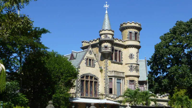 Kamnita stavba Stollmeyer’s Castle s koničastim stolpom in okroglim stolpičem med drevesi v mestu Port of Spain