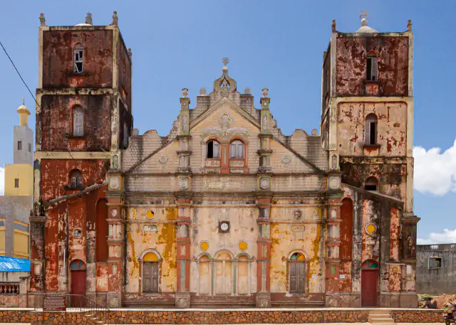 Twin towers and a decorated gable on the red, yellow and green façade of Porto-Novo’s Grande Mosquée under blue sky.