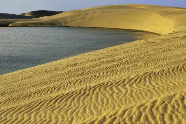 Pale gold sand dunes with fine wind ripples in the foreground curve around a still grey-blue stretch of water, with more dunes rising beyond
