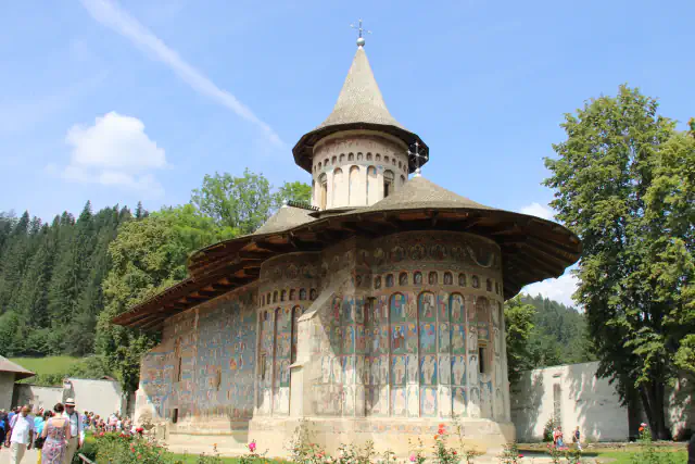 Church of the Voroneț monastery with fresco-covered outer walls and a conical-roofed tower, seen from the courtyard among conifers