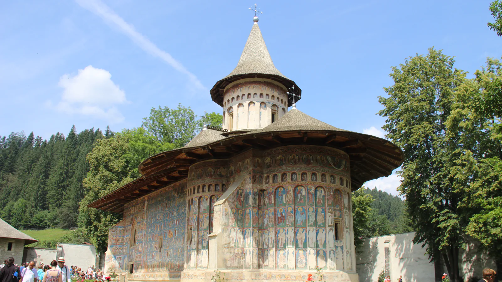 Church of the Voroneț monastery with fresco-covered outer walls and a conical-roofed tower, seen from the courtyard among conifers