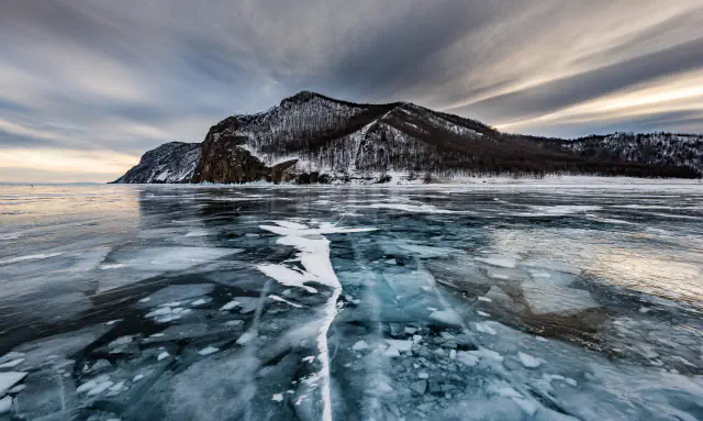 Cracked blue-green ice in the foreground, with a snow-dusted, wooded rocky headland beyond it and streaked clouds above