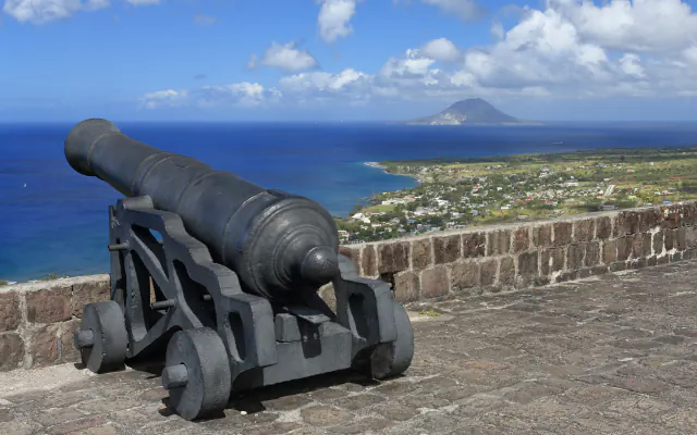 A black iron cannon on a carriage on the stone paving of a fortress rampart, aimed over a low wall towards a deep blue sea, a coastal strip with scattered houses and a distant island with a conical peak