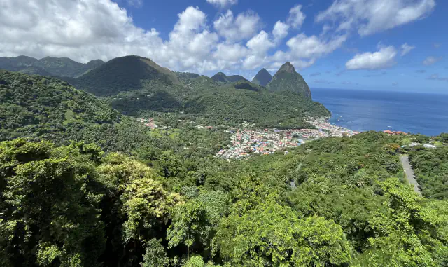Houses of the town of Soufrière beside the sea below forested volcanic hills, with the two pointed Piton peaks rising next to the blue Caribbean under white clouds