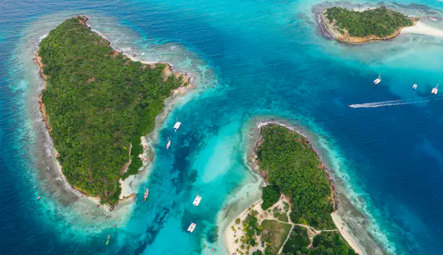 Aerial view of small forested cays in the Tobago Cays, edged with pale sand and turquoise shallows, with sailing boats at anchor and a boat’s wake in deeper blue water