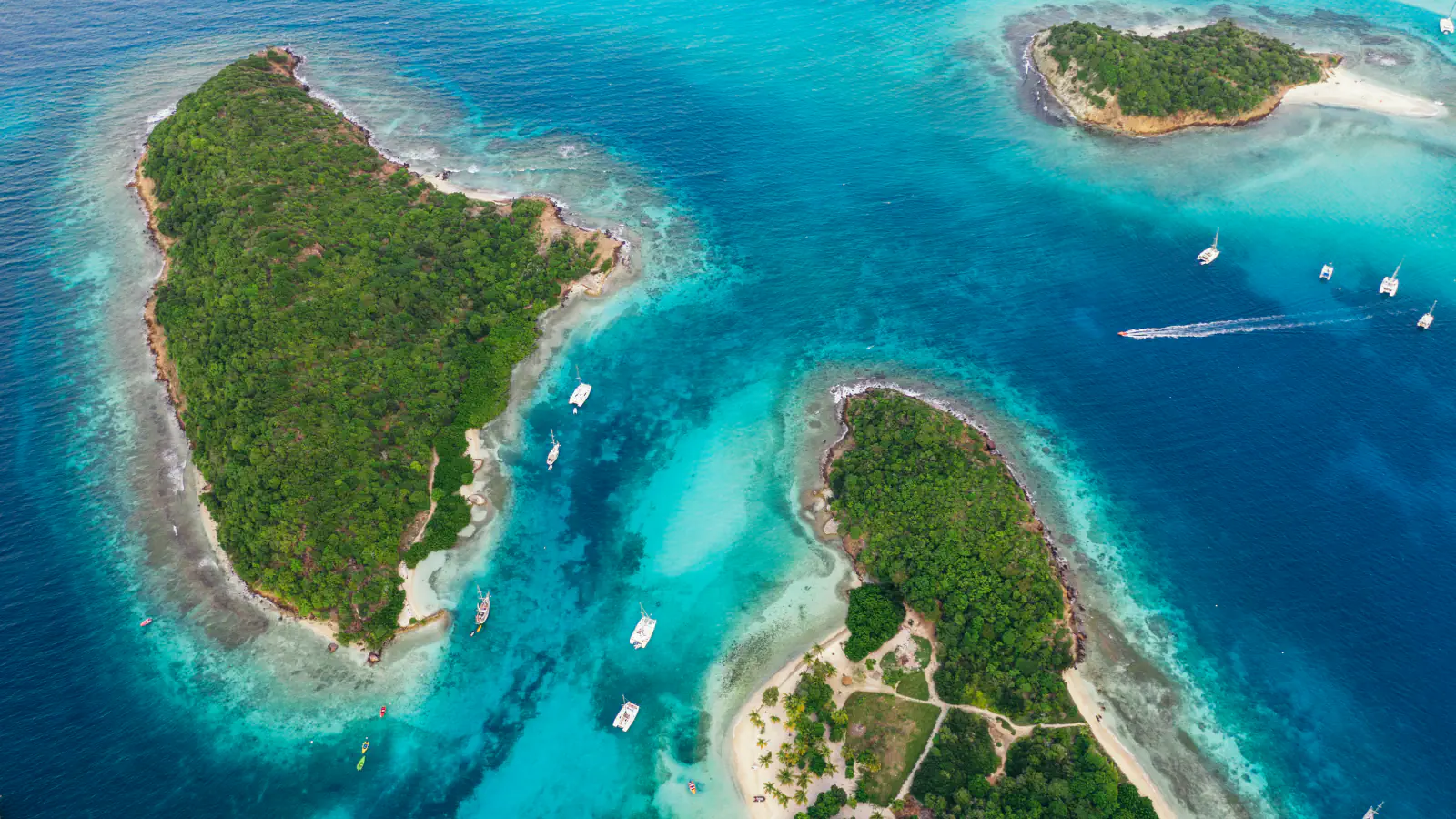 Zračni posnetek majhnih gozdnatih otočkov skupine Tobago Cays, obrobljenih s svetlim peskom in turkiznimi plitvinami, z jadrnicami na sidrišču in sledjo čolna v globlji modrini
