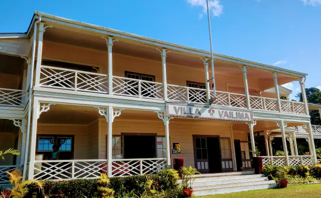 A two-storey cream-coloured wooden house with white lattice verandah railings and a sign reading Villa Vailima above the central entrance, with colourful shrubs along its base, forest at the right and a clear blue sky