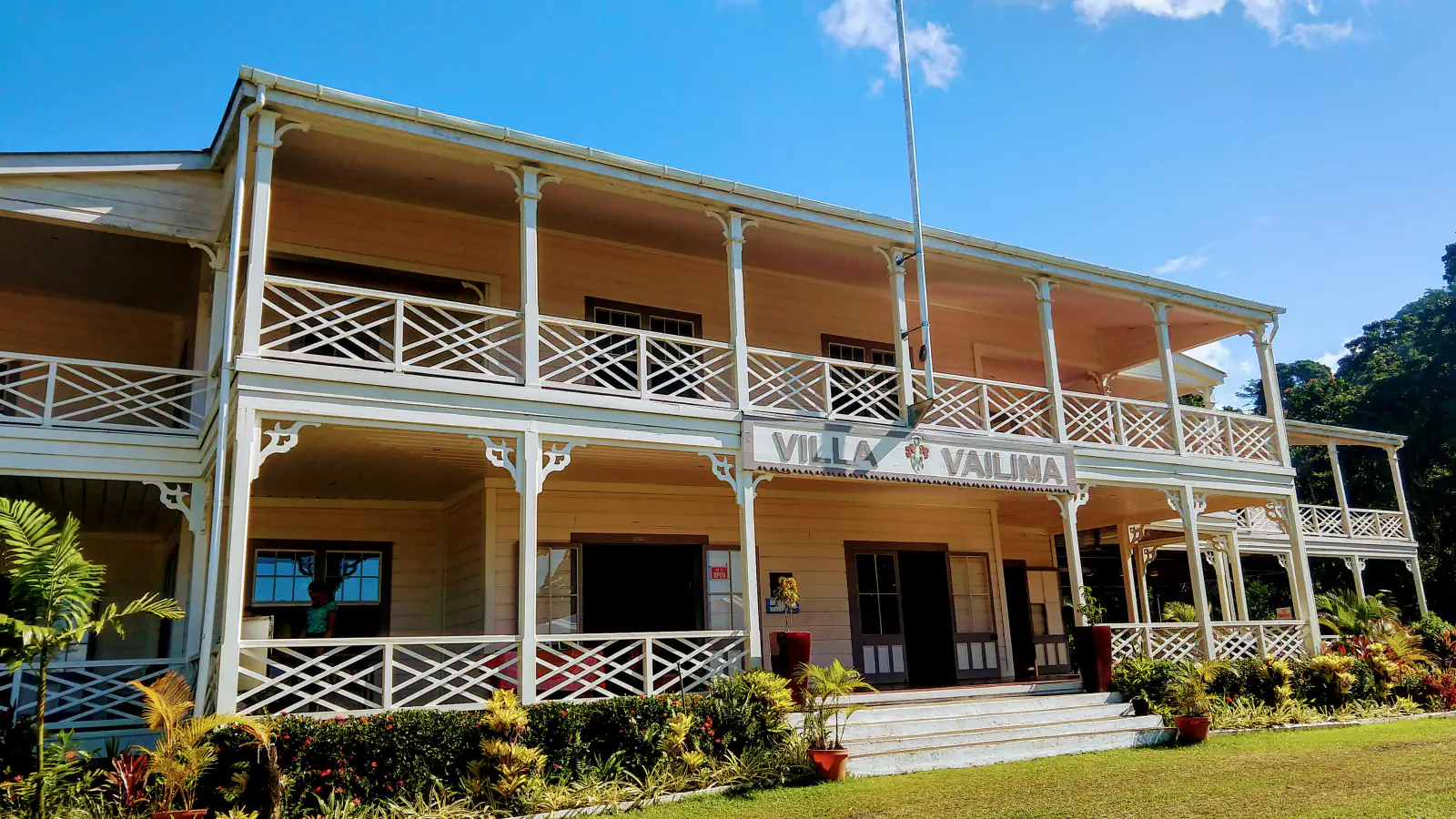 A two-storey cream-coloured wooden house with white lattice verandah railings and a sign reading Villa Vailima above the central entrance, with colourful shrubs along its base, forest at the right and a clear blue sky