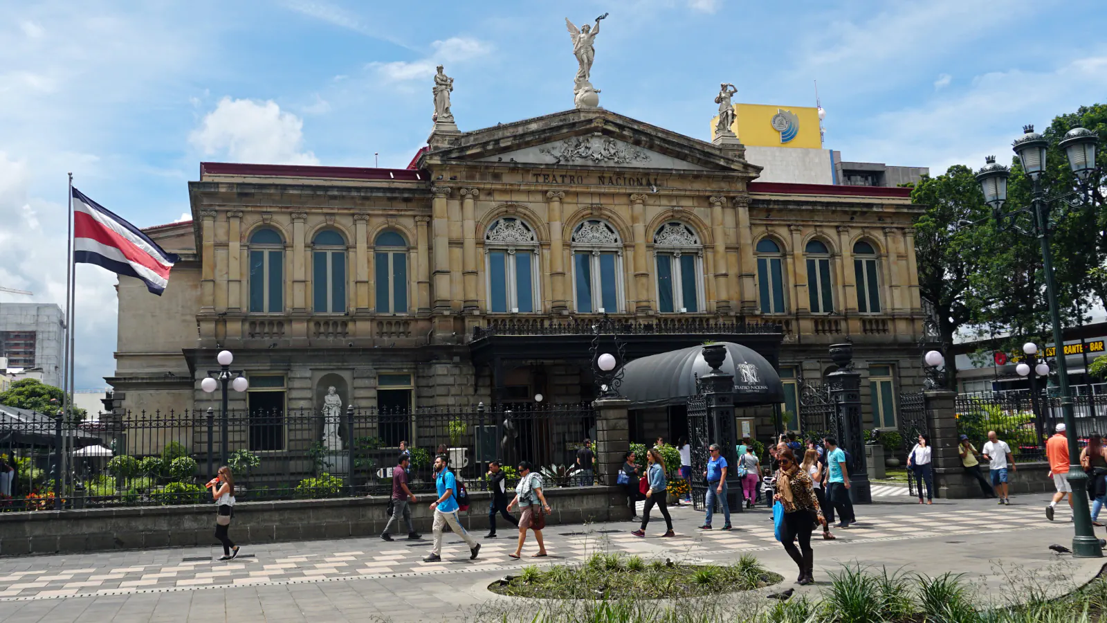 Front of the Teatro Nacional in San José: a sandstone-coloured façade with arched windows and a pediment topped by statues, behind an iron fence, with the Costa Rican flag at left and passers-by on the paved square