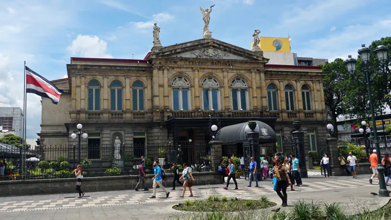 Pročelje gledališča Teatro Nacional v mestu San José: pročelje peščene barve z obokanimi okni in čelnim zatrepom s kipi, za železno ograjo, na levi kostariška zastava in mimoidoči ljudje na tlakovanem trgu