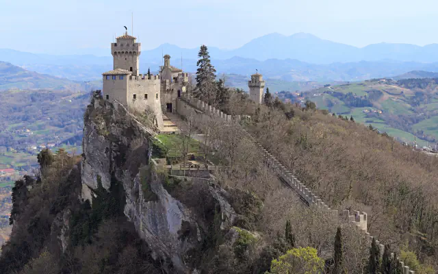 Cesta fortress stands on Monte Titano’s wooded ridge; a crenellated wall descends the slope toward a smaller tower, with hazy hills beyond.