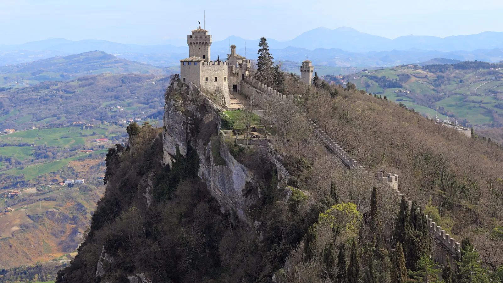 Cesta fortress stands on Monte Titano’s wooded ridge; a crenellated wall descends the slope toward a smaller tower, with hazy hills beyond.