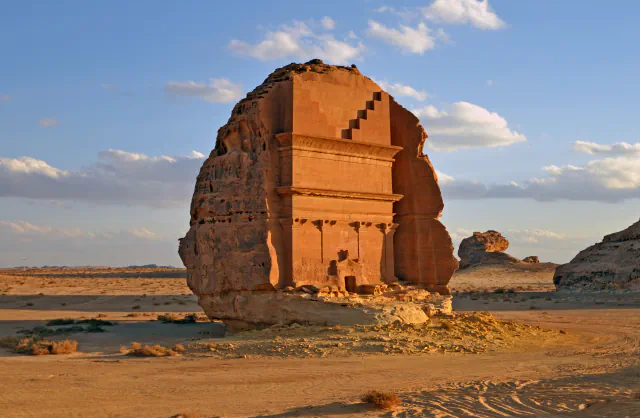 Tomb IGN110 at Hegra, a tall sandstone outcrop standing alone on a sandy desert floor, with a tomb facade carved into its face: stepped blocks at the top, two ledges and four pilasters above a small doorway, under a blue sky with clouds