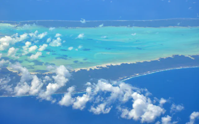 Aerial view of two strips of low, dark green land with white surf along the outer reef, enclosing a pale turquoise lagoon dotted with clouds, with deep blue sea beyond