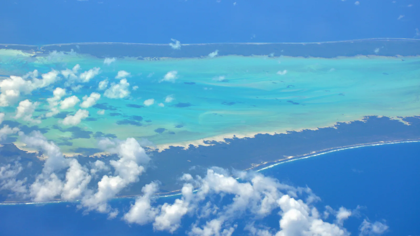 Aerial view of two strips of low, dark green land with white surf along the outer reef, enclosing a pale turquoise lagoon dotted with clouds, with deep blue sea beyond