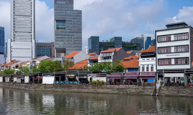 Rows of low shophouses with orange tile roofs beside the Singapore River at Boat Quay, with office towers behind and a stepped stone riverbank in front