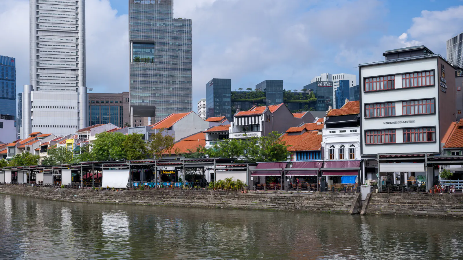 Rows of low shophouses with orange tile roofs beside the Singapore River at Boat Quay, with office towers behind and a stepped stone riverbank in front