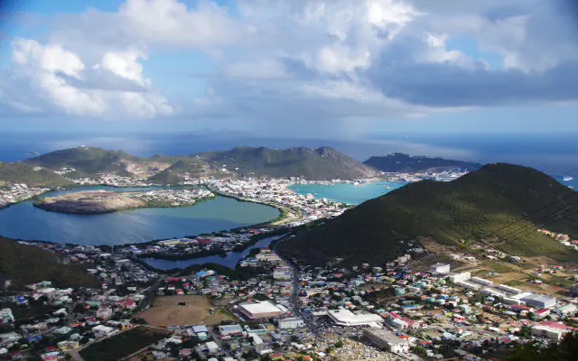 A view from a mountain down on a lagoon with a flat-topped peninsula beside a turquoise bay, a town on the land between them, green hills behind and a cloudy sky