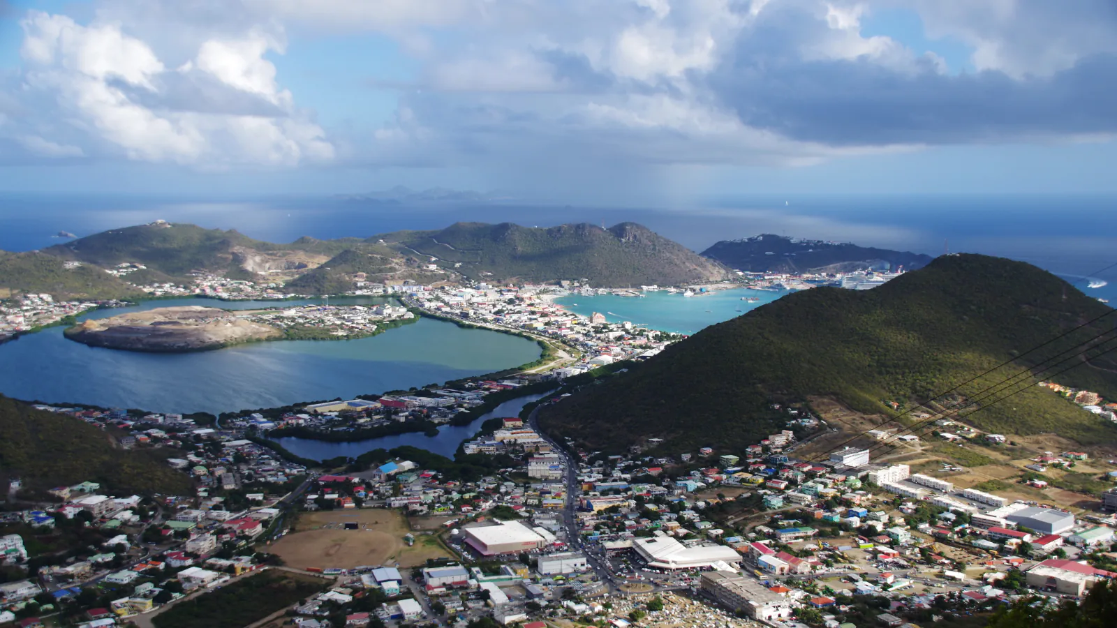 A view from a mountain down on a lagoon with a flat-topped peninsula beside a turquoise bay, a town on the land between them, green hills behind and a cloudy sky