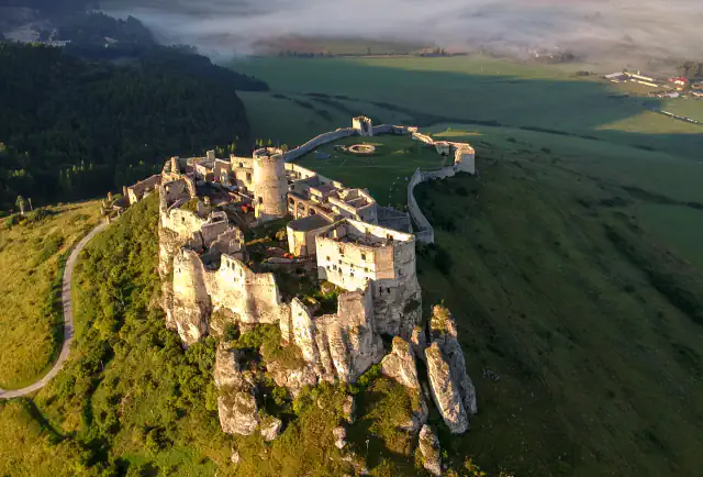 Zračni pogled na Spiški grad na skalnem grebenu v topli jutranji svetlobi; vidni so obzidano dvorišče, zelena polja in meglica nad dolino.