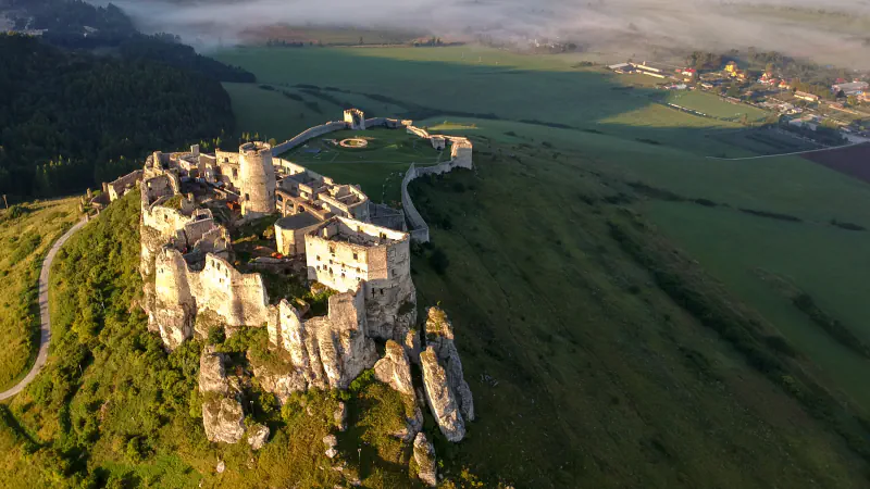 Zračni pogled na Spiški grad na skalnem grebenu v topli jutranji svetlobi; vidni so obzidano dvorišče, zelena polja in meglica nad dolino.