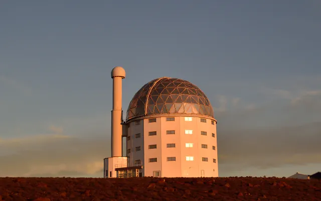 A white round observatory building with a latticed dome and a slim tower topped by a white sphere, lit orange by low sun above red stony ground