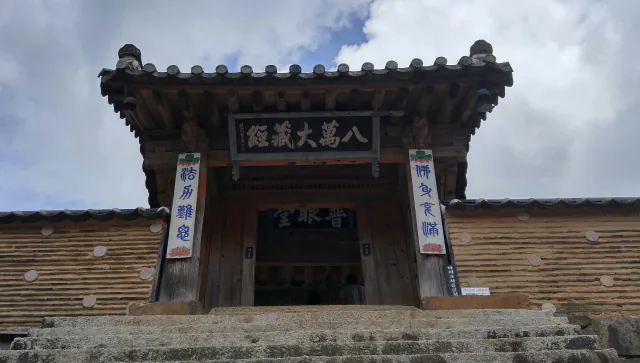 Wooden entrance gate of Haeinsa’s Janggyeong Panjeon seen from the stone steps below, with a signboard in Chinese characters beneath the tiled roof and patterned walls on both sides