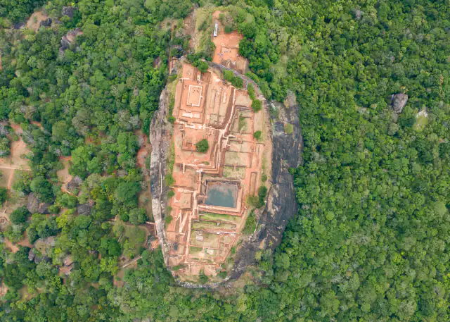 Aerial view straight down on Sigiriya: a flat oval summit of reddish ruined foundations with a rectangular pool, ringed by sheer rock and dense green forest, with cleared plots and paths among the trees on the left