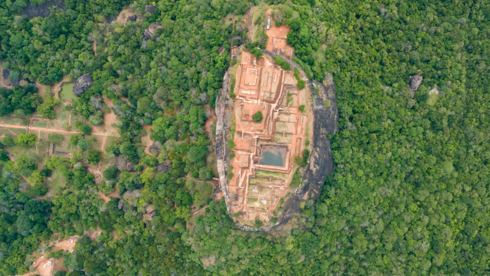 Aerial view straight down on Sigiriya: a flat oval summit of reddish ruined foundations with a rectangular pool, ringed by sheer rock and dense green forest, with cleared plots and paths among the trees on the left