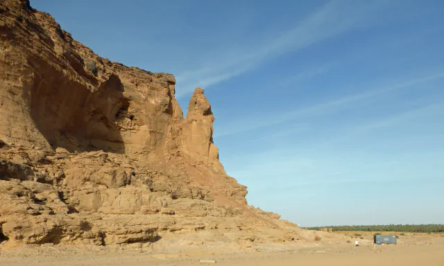 A tall cliff of orange-brown sandstone at the left with a narrow pinnacle standing at its edge; flat sand in front, a dark-green line of palms and a small grey cabin in the distance, and blue sky with thin streaks of cloud.