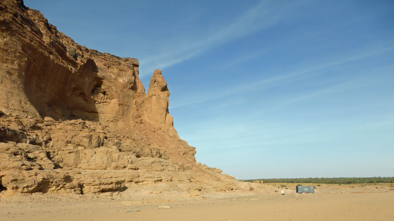 A tall cliff of orange-brown sandstone at the left with a narrow pinnacle standing at its edge; flat sand in front, a dark-green line of palms and a small grey cabin in the distance, and blue sky with thin streaks of cloud.