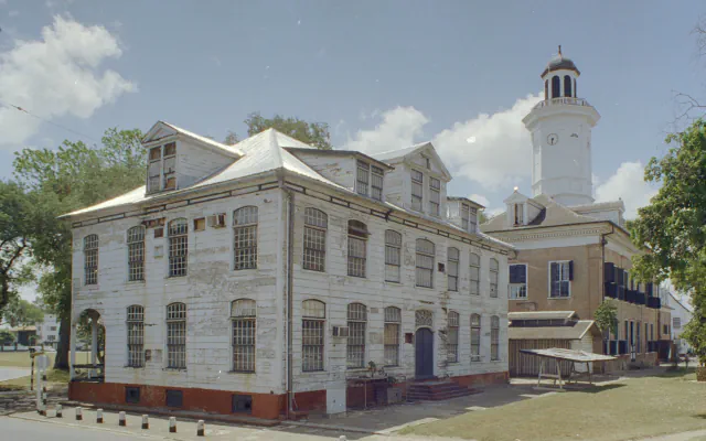 Two-storey wooden building with weathered white boards, arched windows, dormers and a red-brick base, beside a brick building with a white clock tower, on a grassy square under a blue sky