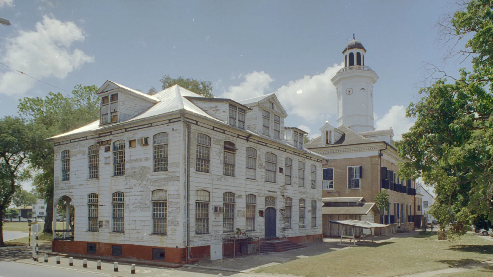 Two-storey wooden building with weathered white boards, arched windows, dormers and a red-brick base, beside a brick building with a white clock tower, on a grassy square under a blue sky