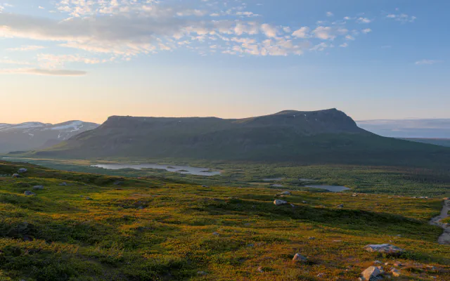 Flat-topped mountain rising above a valley of small lakes and forest, seen from a tundra slope with a path at the right edge