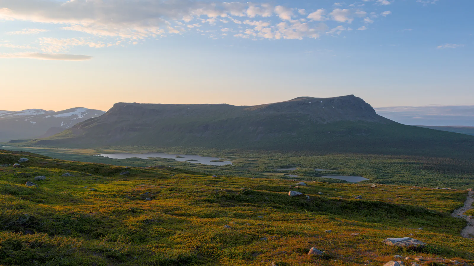Flat-topped mountain rising above a valley of small lakes and forest, seen from a tundra slope with a path at the right edge