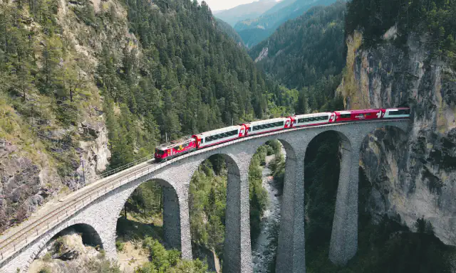 A red-and-white passenger train on the arched stone Landwasser Viaduct above a wooded gorge