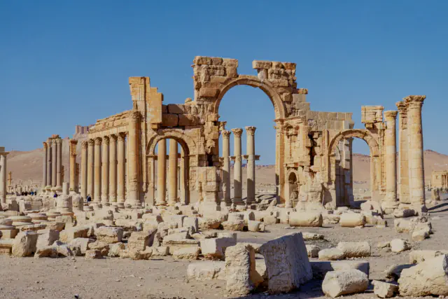 A weathered stone arch with a tall central opening stands beside rows of columns along a colonnaded street, with fallen blocks in the foreground and pale desert hills under a clear blue sky
