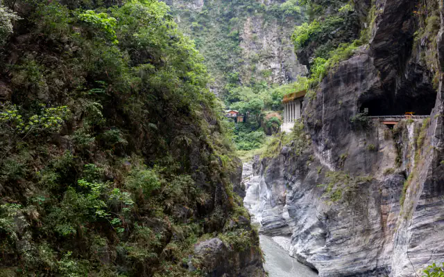 A narrow gorge with steep green slopes on the left and banded grey rock walls on the right, a pale river below, and a small building and tunnel mouth on the far wall