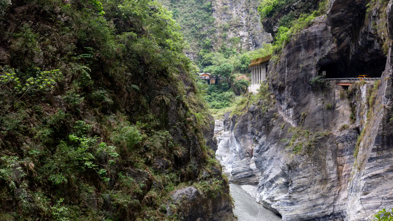 A narrow gorge with steep green slopes on the left and banded grey rock walls on the right, a pale river below, and a small building and tunnel mouth on the far wall