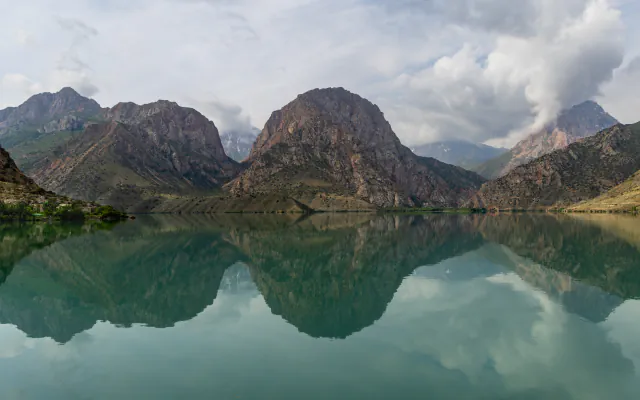 Still, green-tinted lake water mirroring brown rocky peaks and clouds, with trees and a few buildings on the left shore