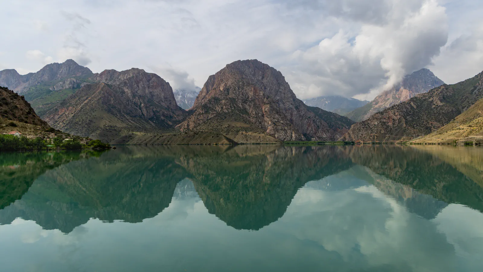 Still, green-tinted lake water mirroring brown rocky peaks and clouds, with trees and a few buildings on the left shore