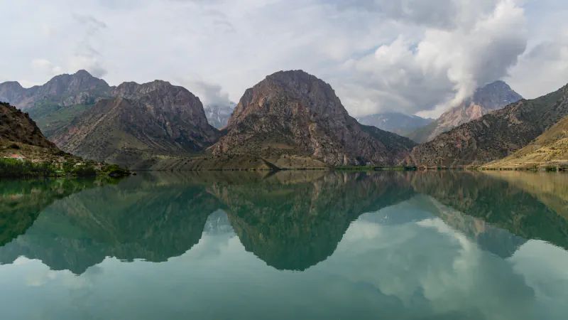 Mirno, zelenkasto jezero, v katerem se zrcalijo rjavi skalnati vrhovi in oblaki, na levem bregu drevesa in nekaj stavb