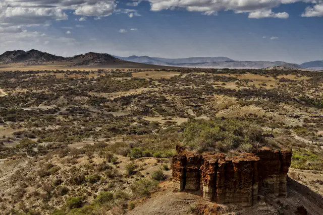 Dry bushland valley with a pale track winding through it, a low dark hill and distant ridges under large clouds, and at lower right a reddish layered rock stack topped with green shrubs