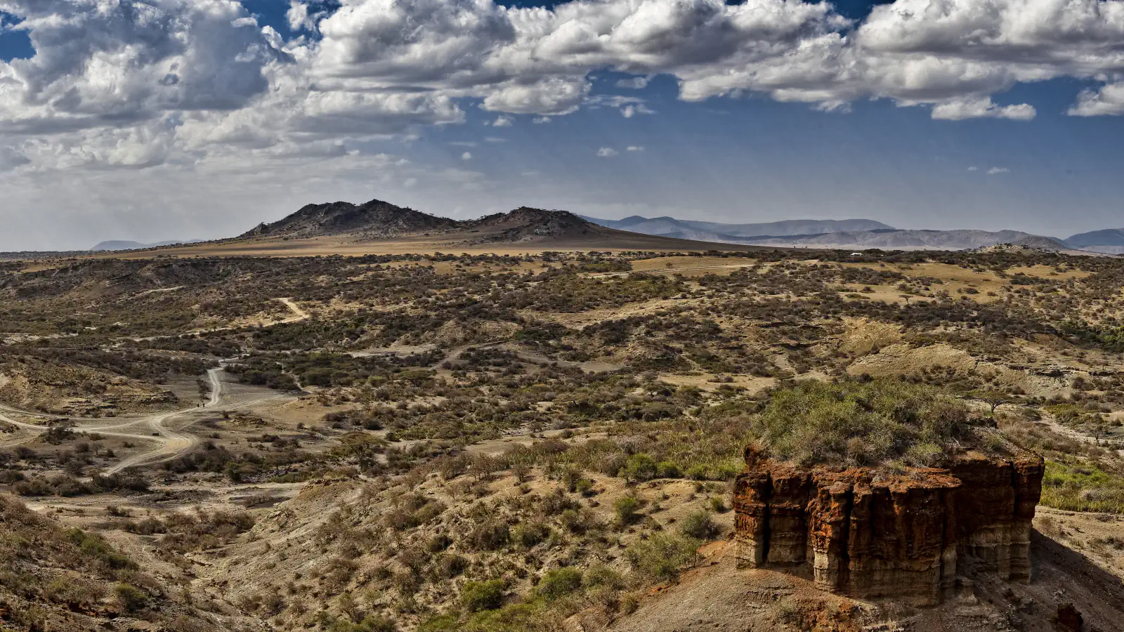 Dry bushland valley with a pale track winding through it, a low dark hill and distant ridges under large clouds, and at lower right a reddish layered rock stack topped with green shrubs
